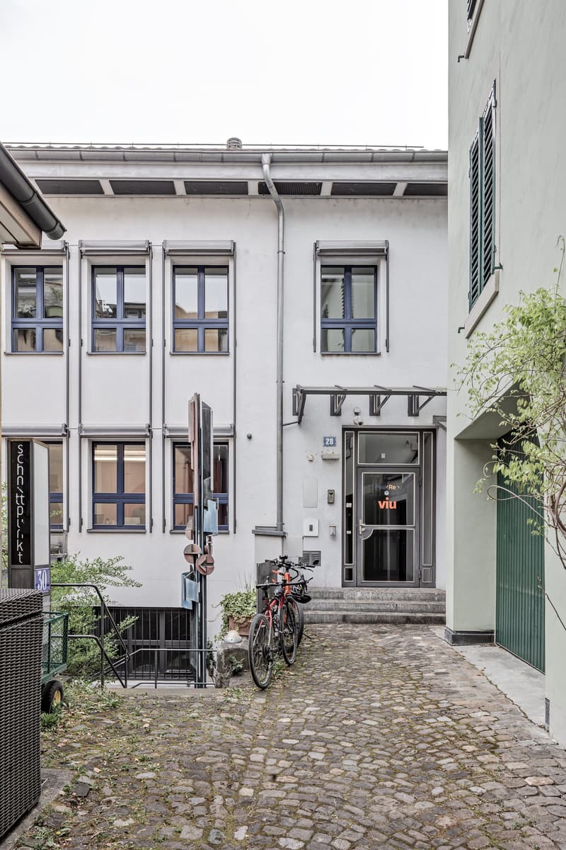 2-story building with white walls, blue-framed windows, and a cobblestone courtyard. There is a bicycle parked in the courtyard and a sign for a business called 'Viu' on the entrance door.