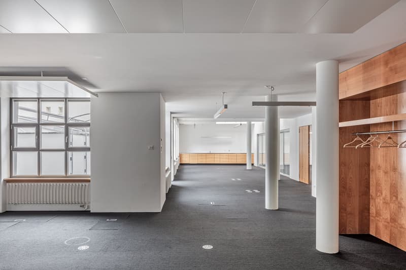 Large open-plan office space with high ceilings, concrete floors, and wooden accents. The space features large windows, pendant lighting, and built-in storage units along the walls.