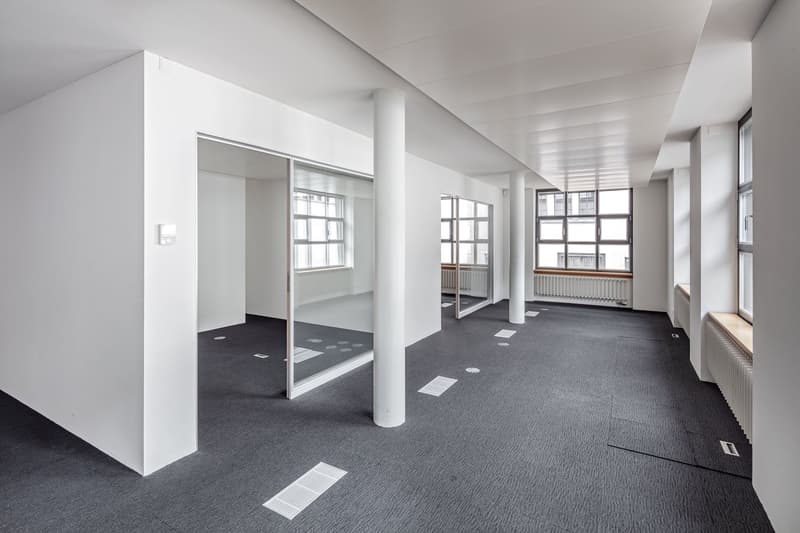 Large open office space with white walls, gray carpeted floor, and several columns. The space has large windows providing natural light. There are sliding glass doors leading to another room or area.