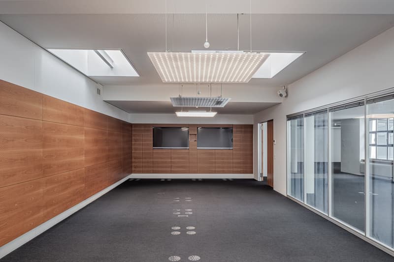 Large open office space with wood paneled walls, recessed lighting, and glass sliding doors. The room has a minimalist and modern design with a dark gray carpet floor.