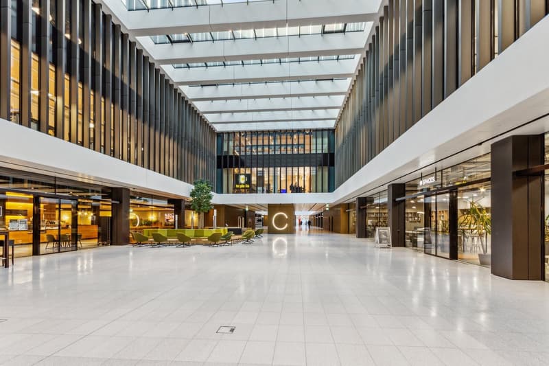 Large open atrium with high glass ceiling, modern design with wooden and glass elements, seating areas, and retail spaces