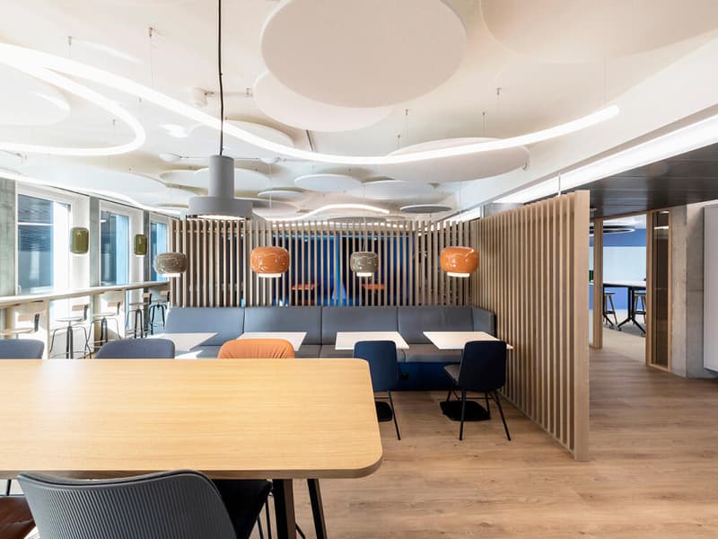 A large open area with wooden flooring, suspended lights, and long tables with chairs. There is a counter and stools in the background. Wooden walls and partitions.