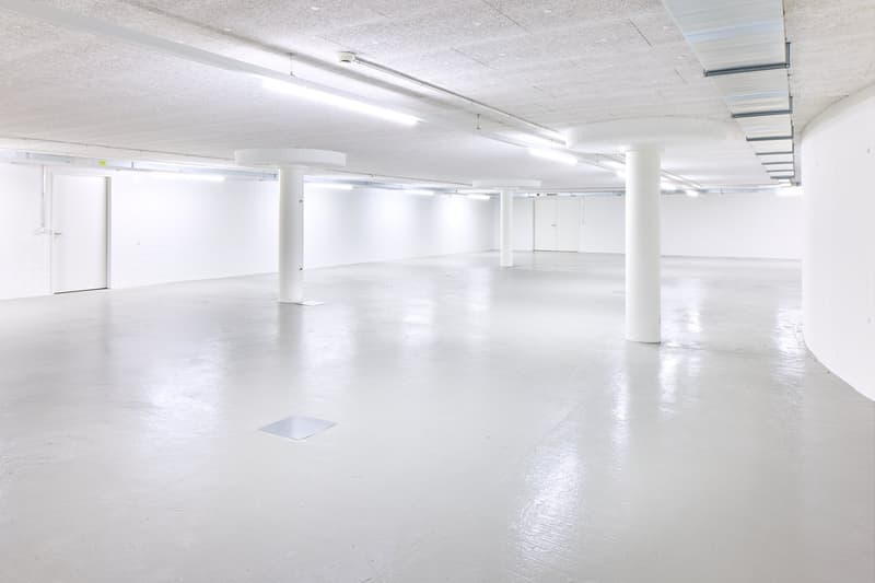 Large open commercial space with white walls, white ceiling, and white concrete floor. The space has multiple columns supporting the ceiling and fluorescent lighting fixtures running along the length of the room. There are no visible features from the pro