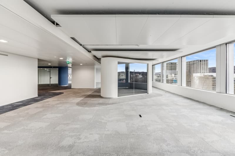 Empty room with carpeted floor, large windows, a central column, exit sign, grey walls, modern design, view of city outside