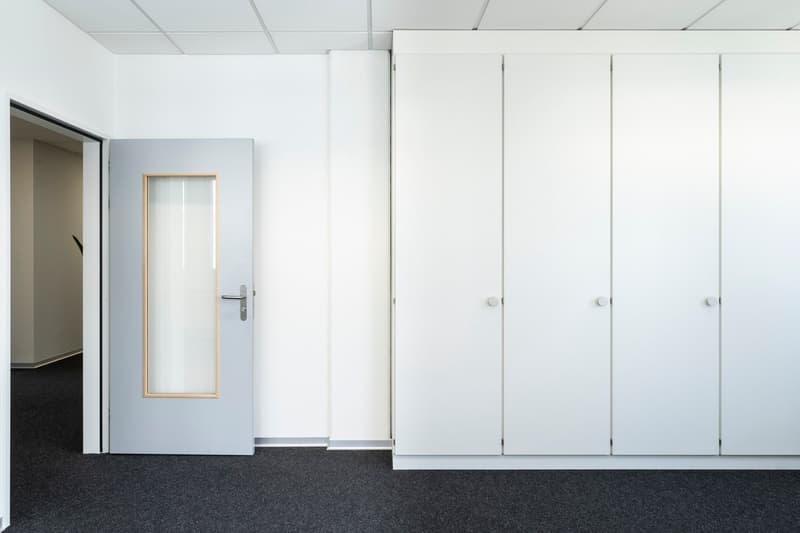 The image shows an office space with white walls, a glass door, and several white cabinets or lockers along the wall. The floor is covered in dark carpeting. The space appears to be a clean and minimalist office environment.