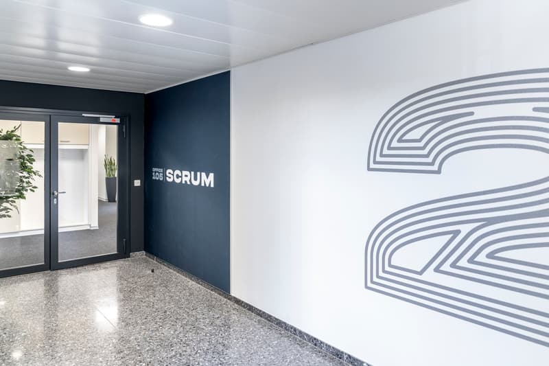 The image shows an office space with a dark blue wall featuring the text 'SCRUM 205' and a large graphic design on the opposite white wall. The room has a glass door entrance, a tiled floor, and some potted plants, indicating a professional and modern off