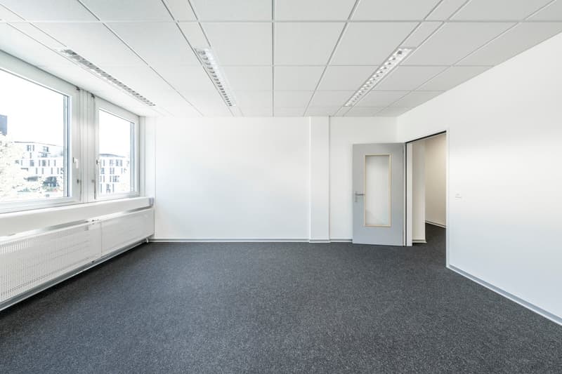 The image shows a large, open office space with white walls, a suspended ceiling with recessed lighting, and a gray carpeted floor. There is a large window overlooking the exterior of the building. The room appears to be empty, ready for use as an office