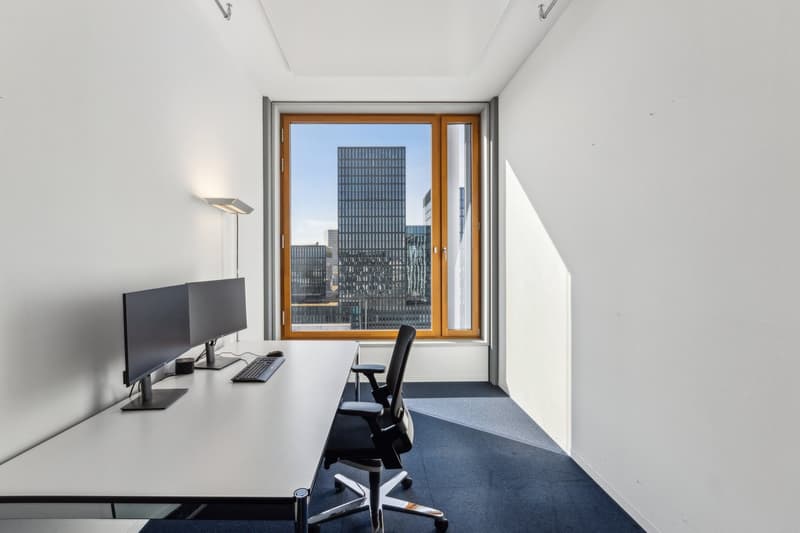 This image shows a modern office space with a large window overlooking a city skyline. The room has a minimalist design with a white desk, a black office chair, and a computer monitor. The floor is covered with a blue carpet, and the walls are painted whi