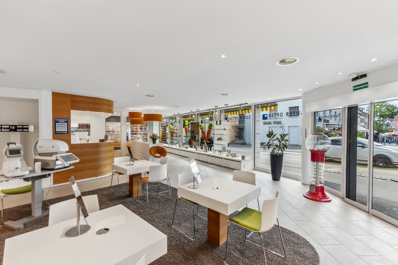 The image shows the interior of a modern retail store or showroom. The space features a clean, minimalist design with white walls, wood accents, and glass display cases. There are various products and displays showcased throughout the room, including what