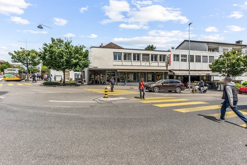 The image shows a busy street scene in an urban area. There are several buildings, including a cafe or shop, and people walking on the sidewalk. The street has yellow crosswalk markings and there are parked cars visible. The sky is partly cloudy.