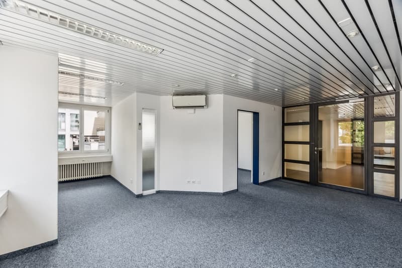 The image shows a large, open office space with a gray carpeted floor, white walls, and a suspended ceiling with recessed lighting. There is a window along one wall and a door leading to another room. The space appears to be set up for office use, with bu