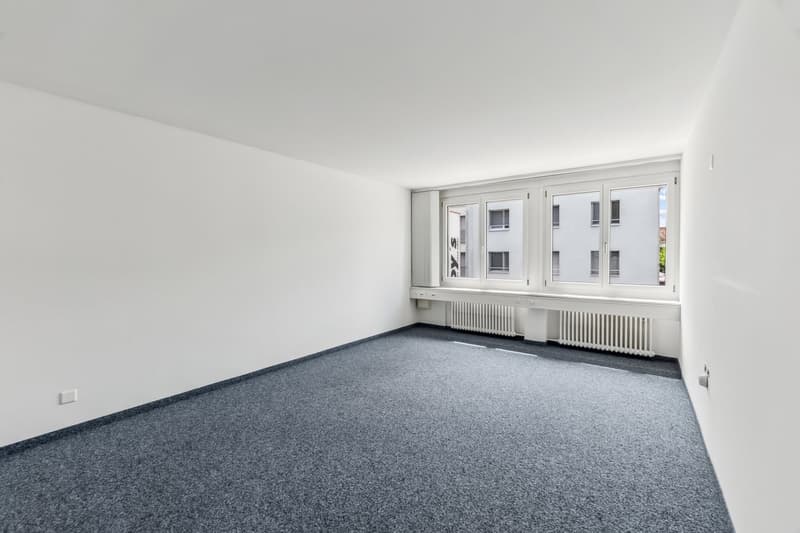 Empty room with white walls, large windows, and gray carpeted floor. The room appears to be a living room or bedroom with no visible furniture or decor.