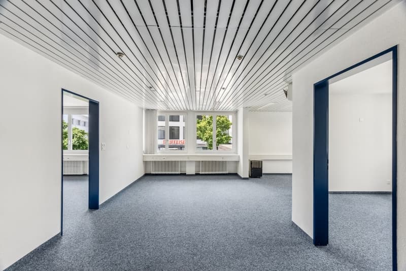 The image shows a large, open office space with a high, slatted ceiling and large windows overlooking greenery outside. The room has a gray carpeted floor and white walls, with dark blue accents around the doorways. The space appears to be empty and ready