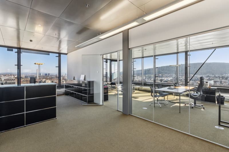 Large open office space with floor-to-ceiling windows offering a panoramic view of the city skyline. The space features a modern, minimalist design with black storage units and a glass-enclosed conference room. The office is equipped with comfortable offi