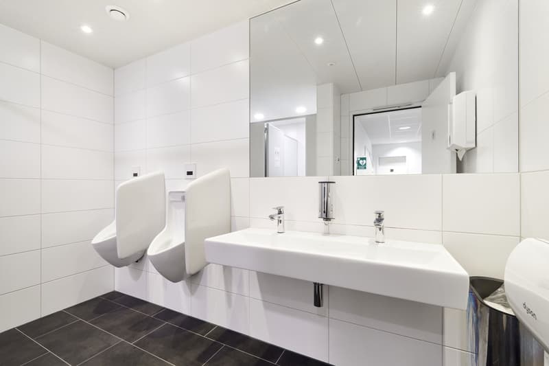 Large, modern bathroom with white tiled walls and floors, two white sinks with chrome faucets, and a dishwasher visible in the corner.
