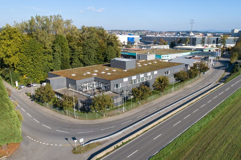 Large commercial building with a green roof, surrounded by trees and a road with multiple lanes. The building appears to be an industrial or office complex with parking spaces visible in the foreground.