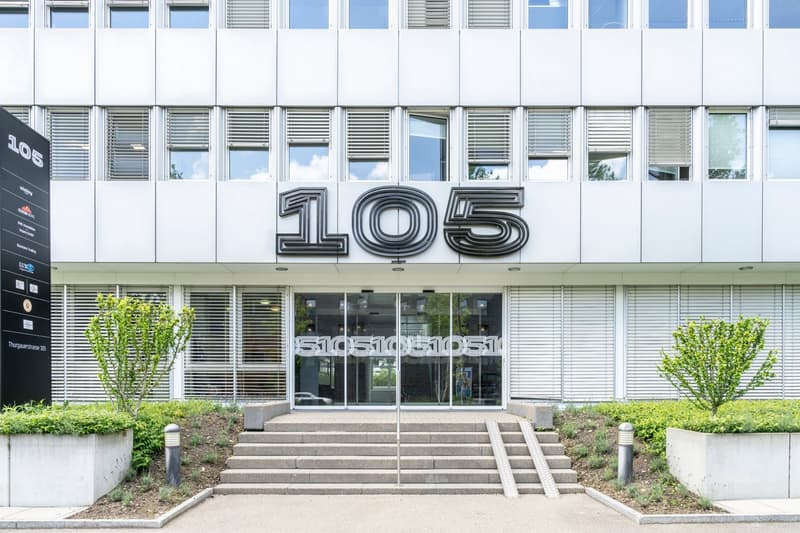 Modern building, glass entrance, number 105 on top, steps leading to entrance, plants on sides