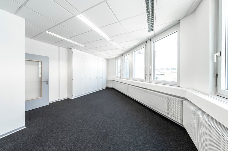 Empty office room, white walls, large windows, radiators, ceiling lights, gray carpet