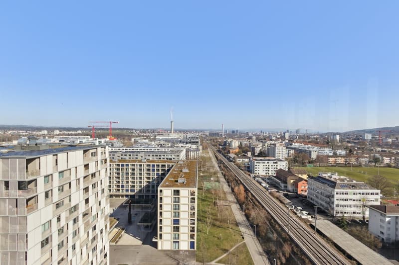Aerial view of urban buildings, train tracks, greenery, and a clear blue sky.