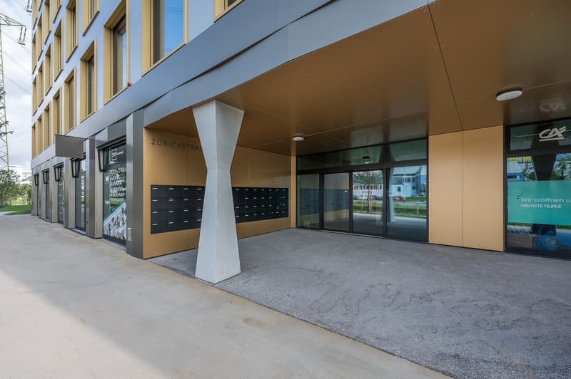 Modern building with glass doors, large windows, and a concrete pillar in front, with a parking area.