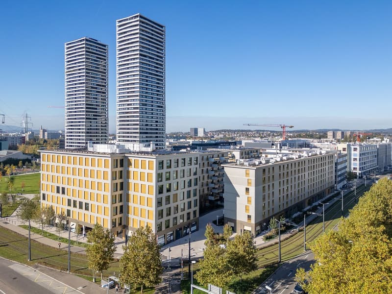 Two high-rise buildings with yellow and white exterior, large windows, and a modern architectural design. The buildings are surrounded by trees and a green lawn. In the distance, there are more buildings and a clear blue sky.