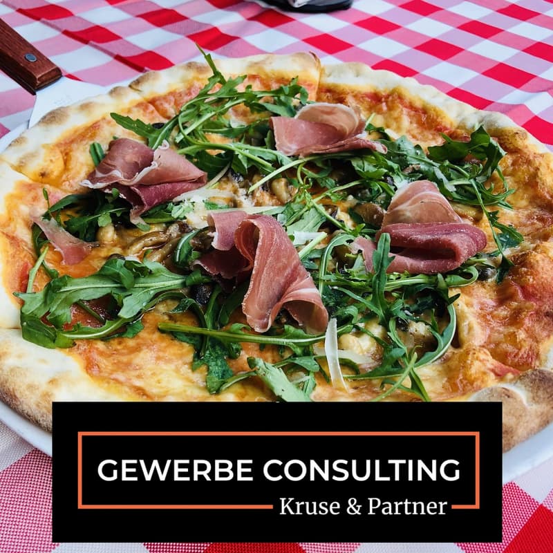 Whole pizza with toppings including prosciutto, cheese, and arugula on a white plate with a red and white checkered tablecloth
