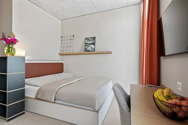 Bedroom with bed, pillows, blanket, a shelf with a picture frame and a grid, TV mounted on the wall, table with fruit bowl, and a vase with flowers