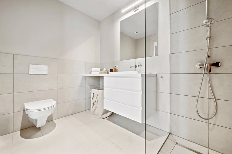 modern clean white tiled bathroom with white vanity, wall mounted toilet, and shower
