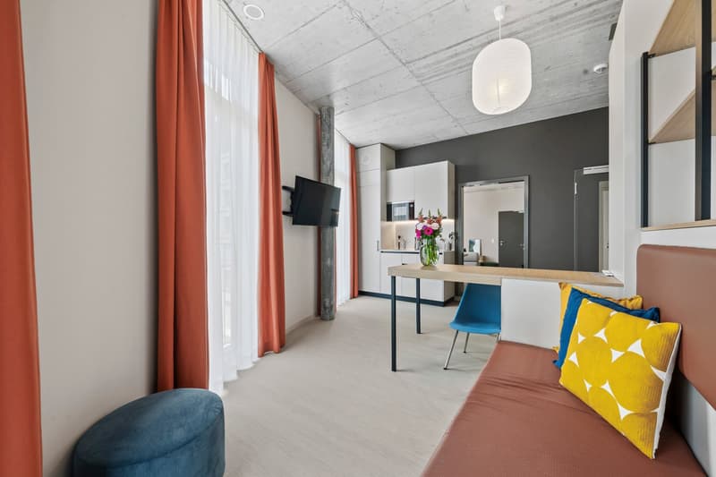 small living area with brown sofa, blue pillow, yellow and white pillow, desk, white chair, white cabinet, TV, open kitchen