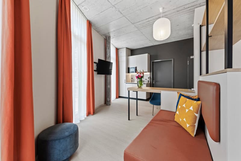 Modern room with wooden flooring, large windows with orange curtains, bed with cushions, bench, desk, blue chair, flat screen TV, microwave, refrigerator, flower vase.