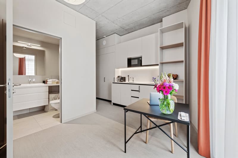 modern open layout, kitchenette, bathroom with toilet and sink, white cabinets, dark tiled floor, white walls, white desk with laptop, chair, white floor tiles