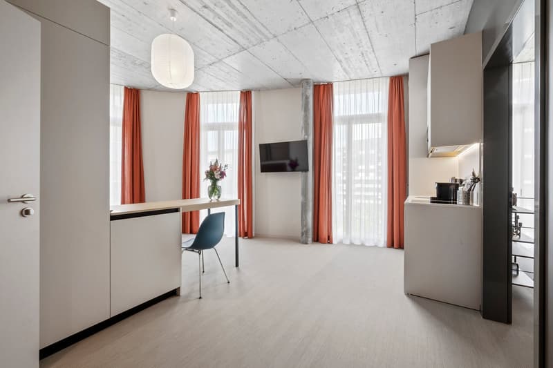 Modern studio apartment with a desk, blue chair, TV, orange curtains, kitchen counter with integrated dishwasher and washing machine, large windows, white ceiling, white walls