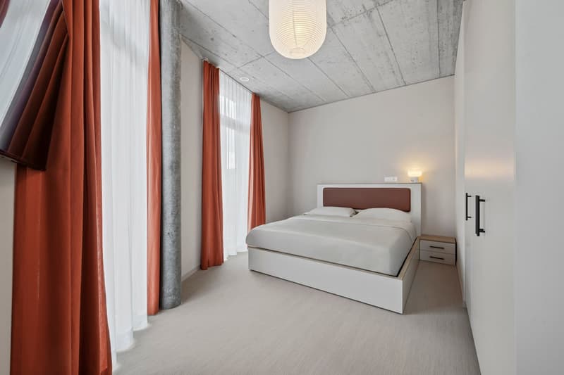 modern bedroom with white walls, ceiling lamp, double bed, wooden nightstand, white carpet, large windows with orange curtains