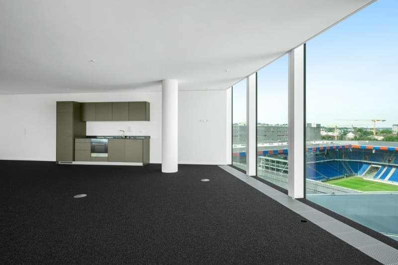Large open-plan living space with a modern kitchen, floor-to-ceiling windows offering a view of a sports stadium, and a spacious, carpeted floor area.
