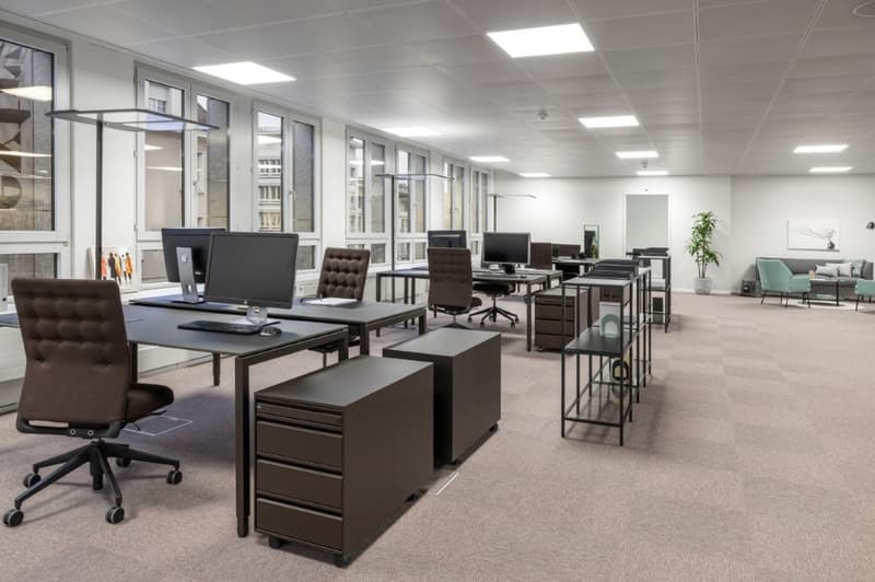 Contemporary office space with desk setups, modern lighting, glass windows, white walls, carpeted floor, and ergonomic chairs.