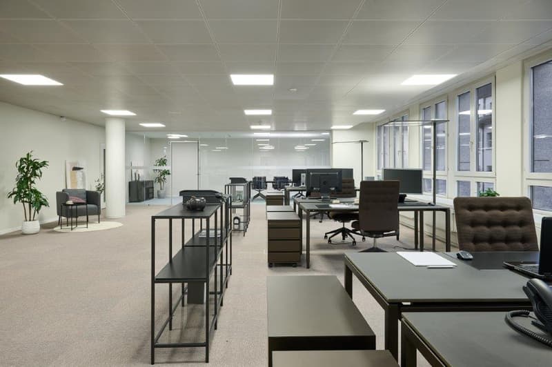 Large open office space with multiple workstations, desks, chairs, and computer equipment. The space has a modern, minimalist design with neutral colors and plenty of natural light from the windows. There are also some potted plants and other decor elemen
