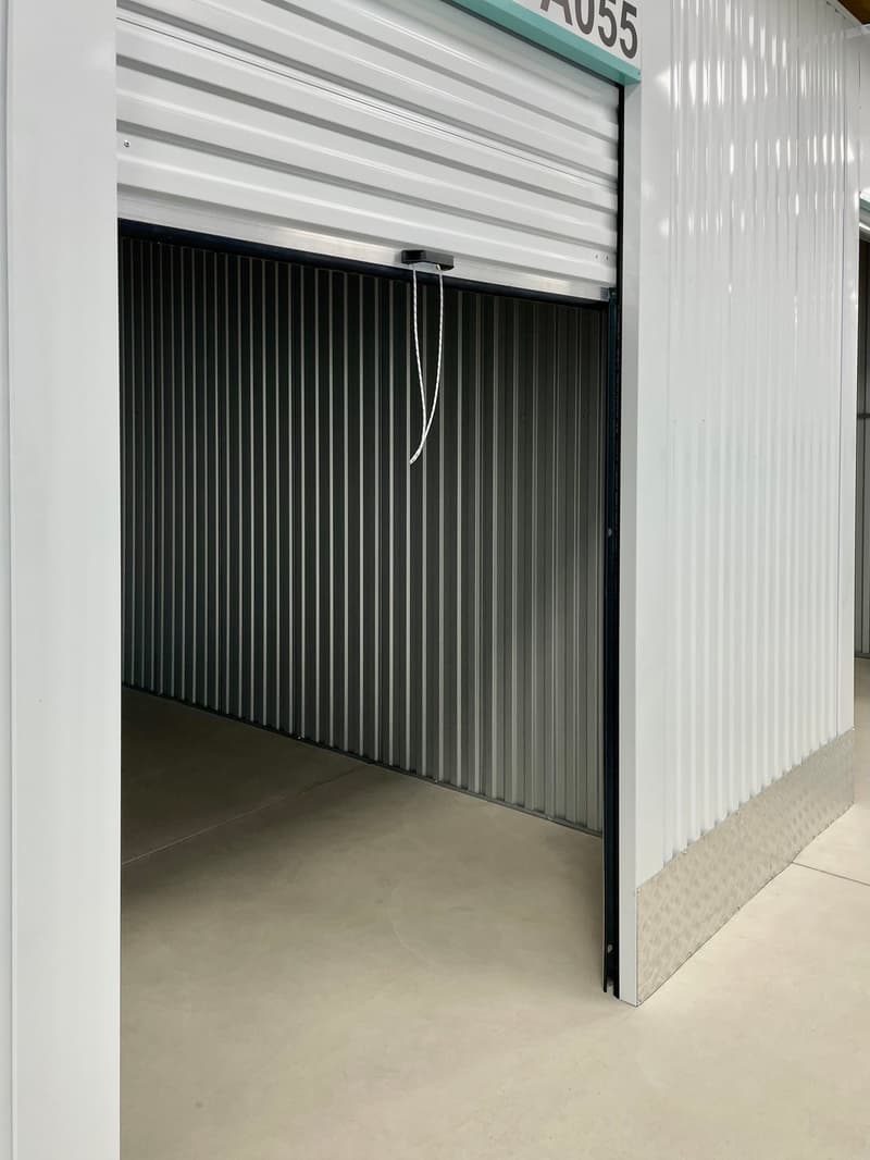The image shows the interior of a storage unit. The unit has a white, corrugated metal exterior and a black, vertical roll-up door. The floor of the unit appears to be a concrete or similar material. The unit is numbered 1055.
