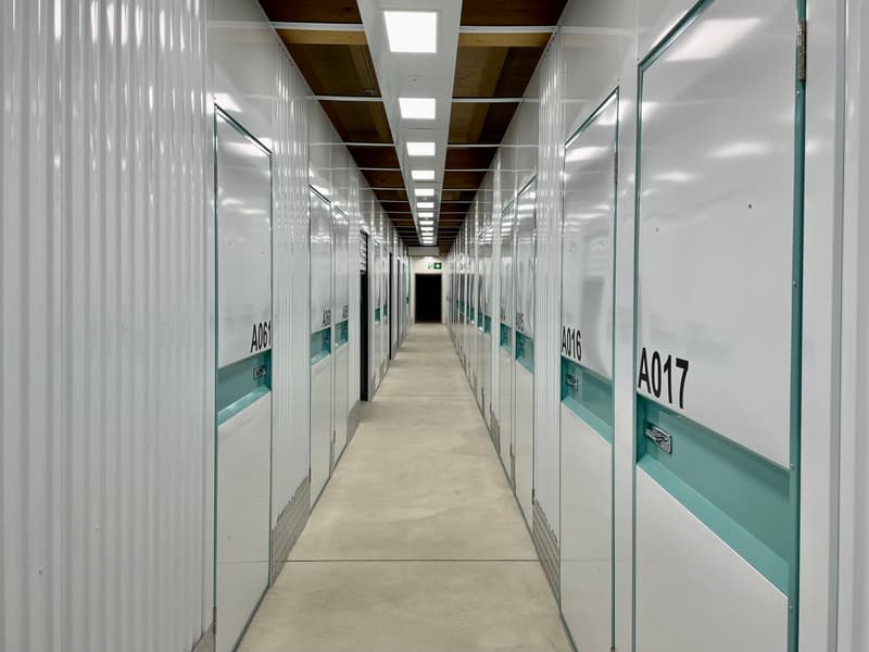 The image shows a long, narrow hallway with multiple storage units or lockers on both sides. The hallway has a concrete floor and the units have glass doors, allowing visibility into the individual spaces. The units are labeled with numbers, indicating th