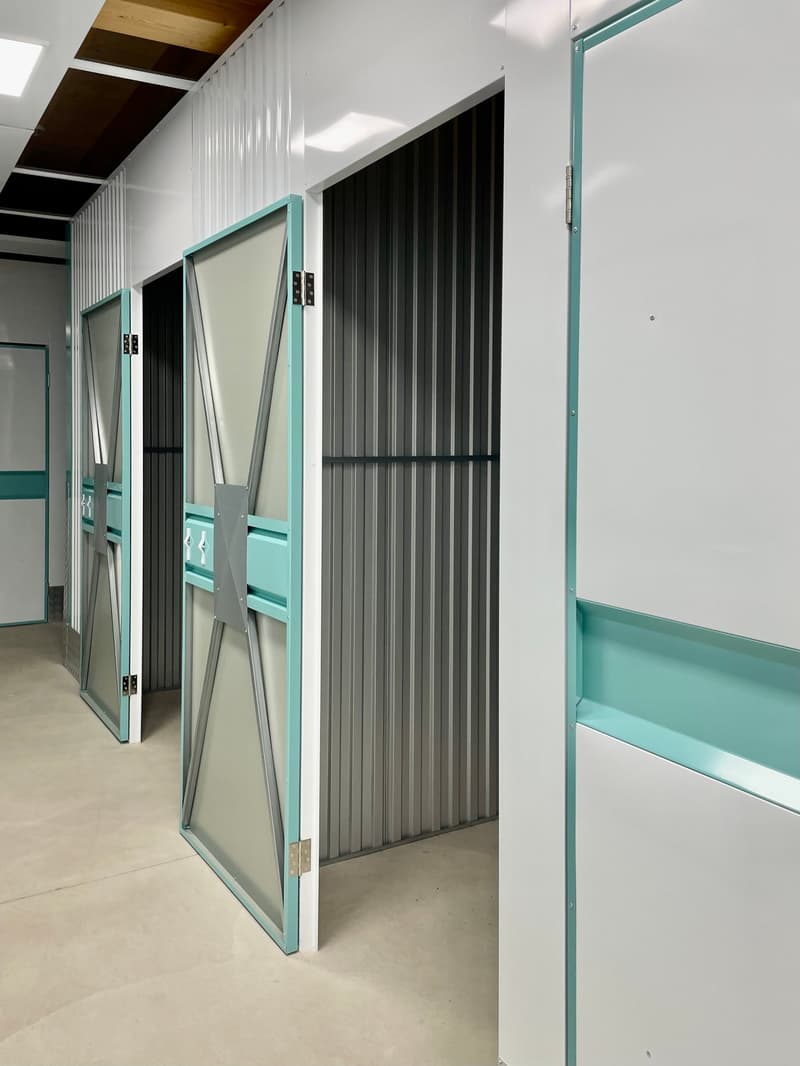The image shows a storage unit with multiple compartments. The compartments have teal-colored frames and sliding doors made of a combination of glass and corrugated metal panels. The walls and ceiling of the storage unit are white, creating a clean and or