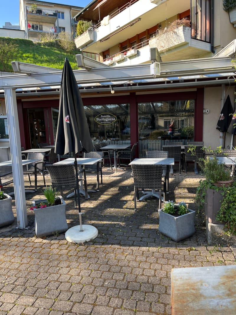 A restaurant's outdoor dining area with brick floors, round tables, and woven chairs. Potted plants and umbrellas add a touch of greenery and shade. Balconies and plants on the upper floors add to the charm. Behind the restaurant, a building's windows and