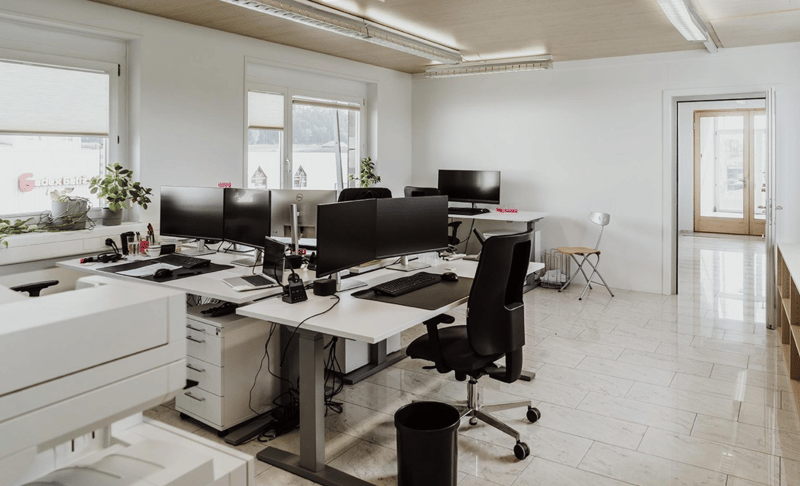 modern office setup with desks, monitors, keyboards, chairs, plants, white walls, windows, and ceiling lights