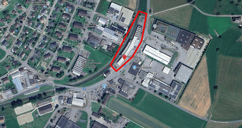 The area features a dense industrial setup with several large buildings, and various facilities, surrounded by fields. Parking lots are visible, with several vehicles. The central focus is an area marked in red, likely indicating a specific plot or point