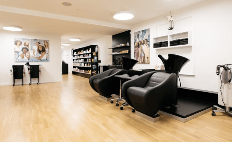 A hair salon interior with wooden floors, white walls, round lamps, black swivel chairs, shampoo trolleys, shelves with products, and framed pictures on the wall.