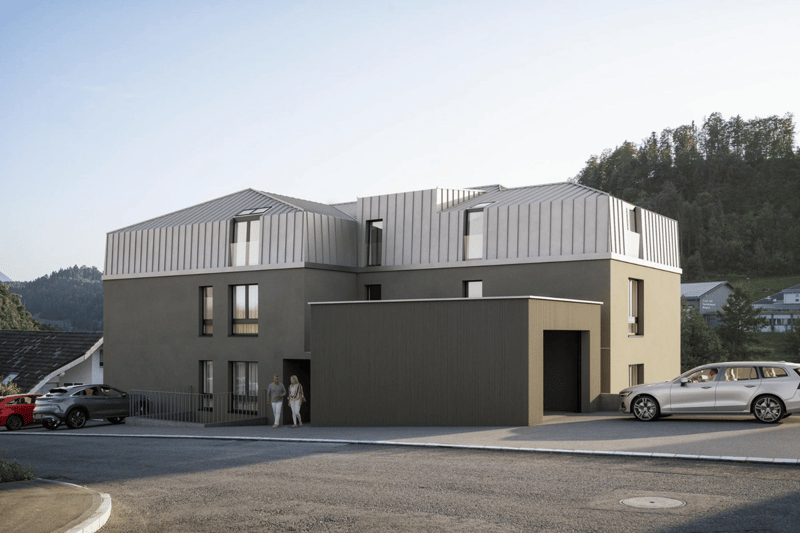 Modern residential building with multiple levels, grey exterior, numerous windows, driveway for cars, entrance with fencing, nearby parked vehicles
