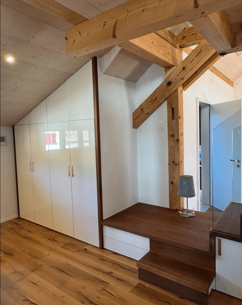 A room with built-in wardrobes, wooden floors, and a bench underneath the slanted ceiling.