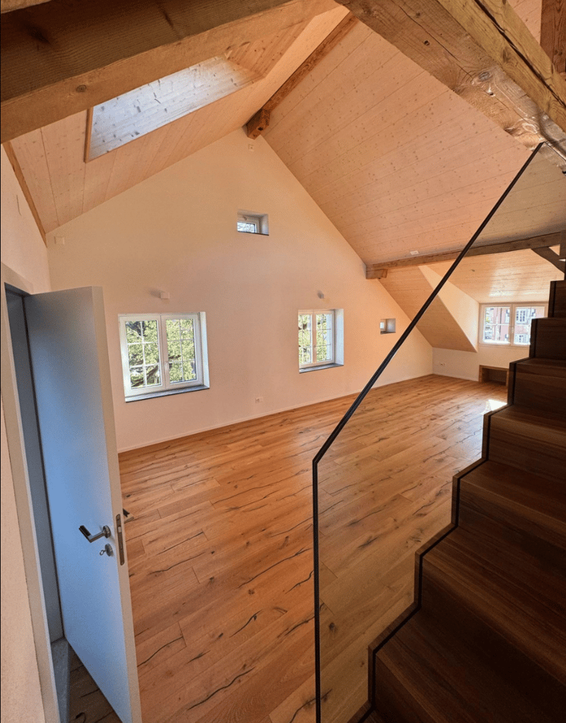 an empty room with wooden floors, open-beamed ceiling, wooden stairs, two small windows
