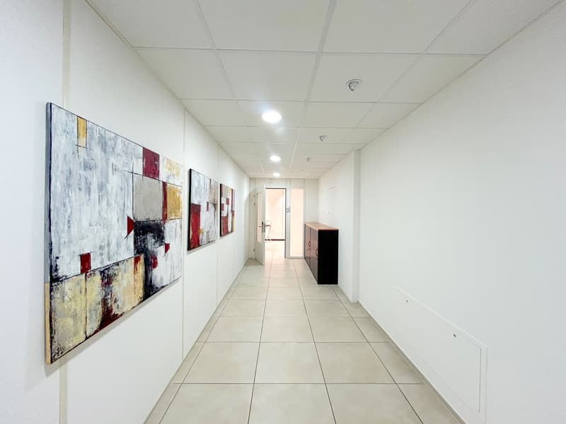 Empty hallway with white walls, white tiles, three paintings, a wooden cabinet, and a door.