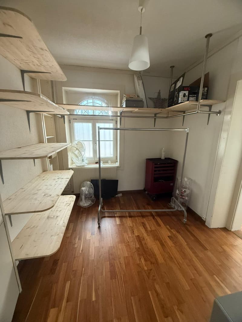 Empty room with wooden flooring, metal clothing rack, wooden shelves, and an arched window