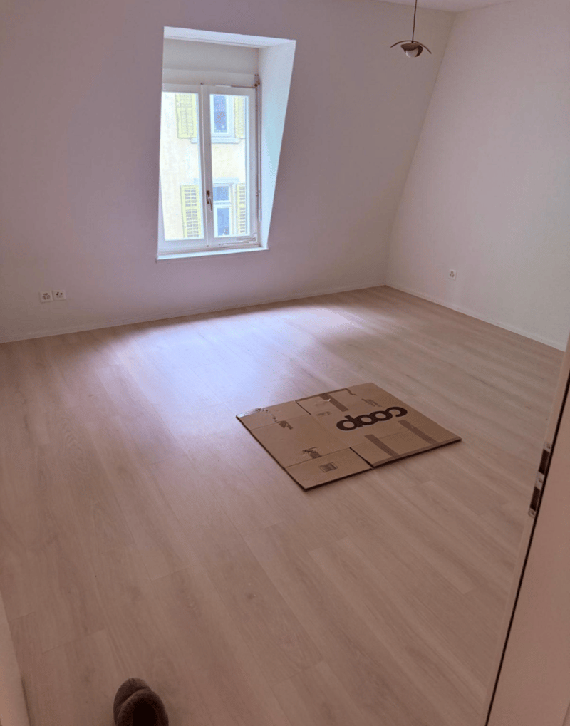 Empty room with white walls, wooden floor, window, cardboard box, and electrical outlets