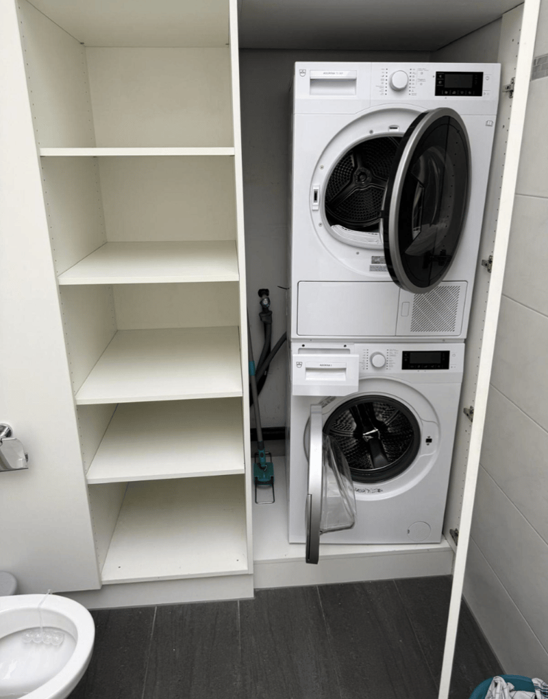 A room with a washing machine and a dishwasher stacked on top of each other, a cabinet, and a toilet.
